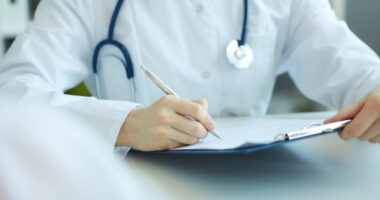 A doctor in a white lab coat with a stethoscope writing on a chart.