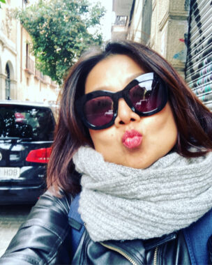 A selfie of a woman wearing stylish dark sunglasses, a gray scarf, and a black leather jacket. She is standing on a city street and blows a kiss to the camera.