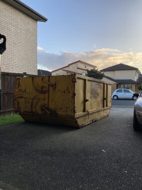 A large yellow dumpster sits in a driveway next to a car in a neighborhood. It appears to be dusk.