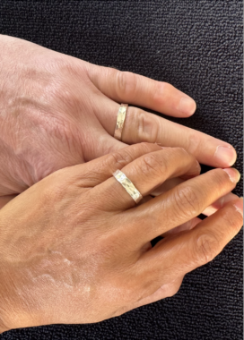 Two left hands, a man's and a woman's, rest together atop a black surface. Both are wearing gold bands on their ring fingers.