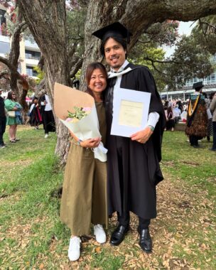 A mother and son embrace under a tree during a college graduation ceremony. The mother is holding a bouquet of flowers while the son, dressed in cap and gown, holds his diploma.