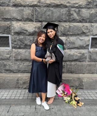 A woman in a dark dress and white shoes stands in front a stone building aside a taller woman in graduation garb. Yellow flowers are on the concrete behind her.